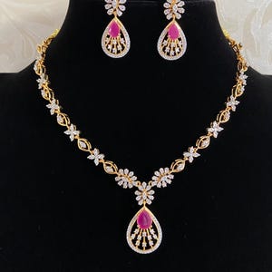May include: A gold-toned necklace and earring set featuring teardrop-shaped pink stones surrounded by clear white crystals. The necklace chain consists of repeating floral-inspired links, while the matching earrings mirror the central pendant design.