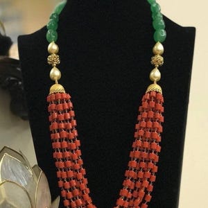 May include: A multi-strand necklace featuring numerous small, cylindrical red beads arranged in a cascading design. The necklace includes gold-toned metal spacers, smooth cream-colored spherical beads, and translucent green beads.