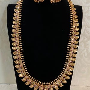 May include: A gold-colored necklace and matching earrings set displayed on a black mannequin. The necklace features a repeating paisley-like design with red and green accents. The earrings are bell-shaped with similar detailing.