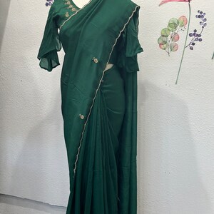 May include: A dark green silk saree with gold trim and embellishments. The saree is draped on a mannequin and has a matching green blouse with ruffled sleeves.