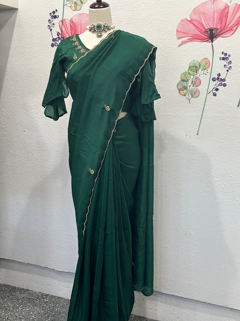 May include: A dark green silk saree with gold trim and embellishments. The saree is draped on a mannequin and has a matching green blouse with ruffled sleeves.