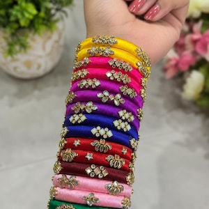 May include: A stack of multi-colored silk thread bangles featuring various shades including yellow, pink, purple, blue, red, maroon, and green. Each bangle is adorned with small, reflective metallic accents and clear stone embellishments.