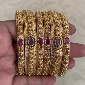 May include: A set of gold-colored bangles with intricate embossed patterns. Each bangle features alternating oval-shaped red and green gemstones. The bangles are held in a hand, showcasing their design and color.