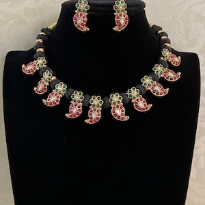 May include: A gold-toned necklace and earring set. The necklace features a black cord with gold beads and paisley-shaped pendants adorned with red and green gemstones. The matching earrings have the same paisley design. The jewelry is displayed on a black mannequin.