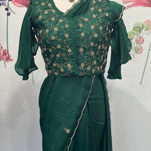 May include: A dark green satin saree with a beaded border and a matching green embroidered blouse with a ruffled sleeve. The blouse has a floral design with gold and white beads.