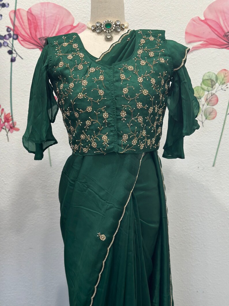 May include: A dark green satin saree with a beaded border and a matching green embroidered blouse with a ruffled sleeve. The blouse has a floral design with gold and white beads.