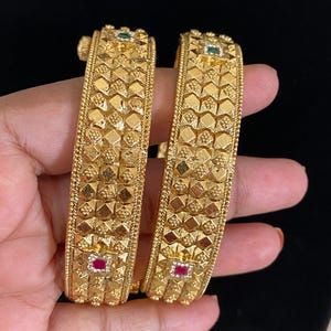 May include: Two gold-toned bangles featuring a textured, geometric pyramid pattern across the surface. Each bangle is accented with small, square-cut green and red stones set at intervals within the intricate, raised metalwork design.