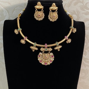 May include: A gold-toned necklace and matching earring set featuring intricate metalwork, small green and pink stones, and a central pendant with a fan-shaped design. The necklace has a rigid circular band adorned with dangling decorative charms.