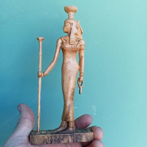 Peut inclure: Une figurine en bois de la déesse égyptienne Nephthys, debout avec un bâton dans sa main gauche et un ankh dans sa main droite. La figurine est peinte en brun clair et a une base avec le mot "NEPHTHYS" inscrit dessus.