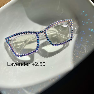 May include: Clear plastic framed glasses with rhinestones around the frame. The glasses have a +2.50 prescription and are labeled "Lavender".