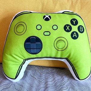 May include: A lime green plush pillow shaped like an Xbox controller. The pillow has black buttons labeled 'X', 'Y', 'A', and 'B'.