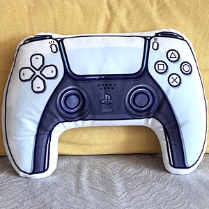 Può includere: Un cuscino a forma di controller PlayStation 5, bianco e nero. Il cuscino è realizzato in tessuto morbido e ha un design realistico.