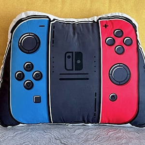 May include: A white pillow shaped like a Nintendo Switch controller. The left side is blue with a directional pad and buttons. The right side is red with a directional pad and buttons. The middle section is black with the Nintendo Switch logo.