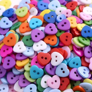 May include: A large assortment of heart-shaped buttons in various colours, including red, blue, green, purple, pink, yellow, and white. The buttons have two holes each.