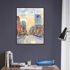 May include: A framed watercolor painting depicting a city street scene at sunset. The artwork features tall buildings, glowing streetlights, and vehicles on a wet road, with a warm yellow and orange sky reflecting on the pavement below.