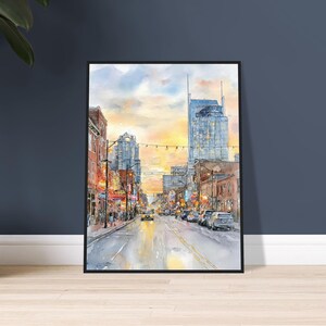 May include: A framed watercolor painting depicting a city street scene at sunset. The composition features tall buildings, glowing street lights, and vehicles on a road. The sky is filled with warm yellow and orange hues, contrasting with the cool blue tones of the architecture and the street level, creating a vibrant urban atmosphere.