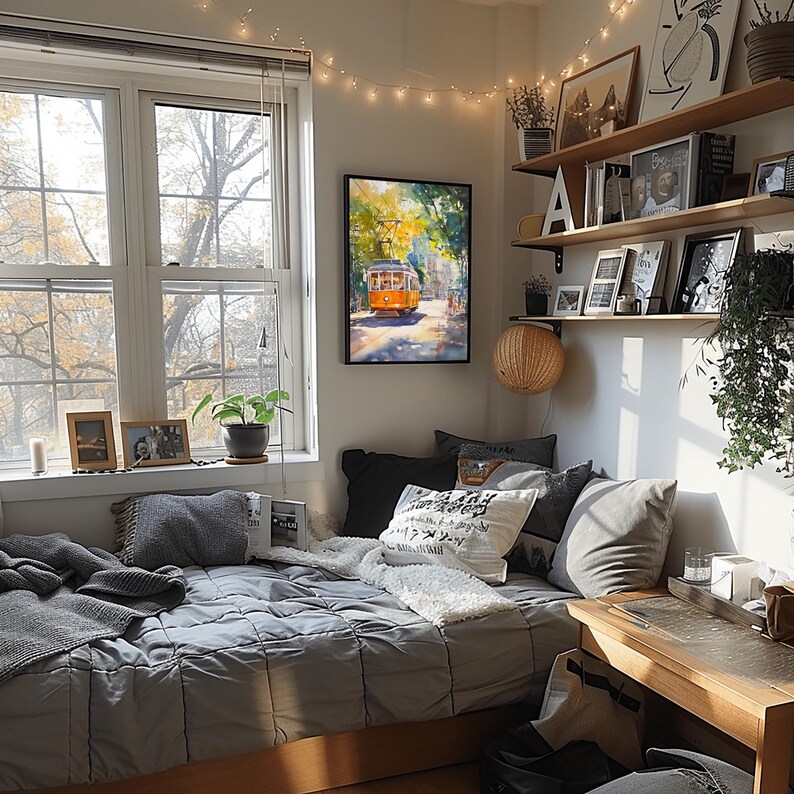 May include: A cozy bedroom with a gray bed, a white throw blanket, and a framed watercolor painting of a yellow trolley car. The room has a window with a view of trees, a small desk, and string lights.