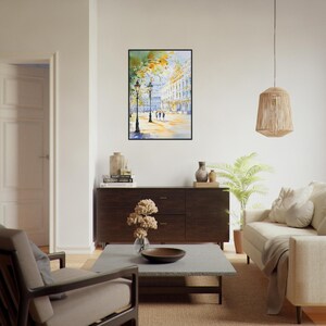Op de afbeelding: Een aquarel schilderij van een stadstraat sc&egrave;ne met gebouwen, bomen en mensen die lopen. Het schilderij is ingelijst in een zwart frame en hangt aan een witte muur.