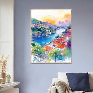 Op de afbeelding: Aquarel schilderij van een stadsgezicht met een brug over een rivier. Het kunstwerk toont gebouwen met oranje daken, palmbomen en een levendige lucht met tinten van oranje en paars. De schildering is ingelijst in licht hout.