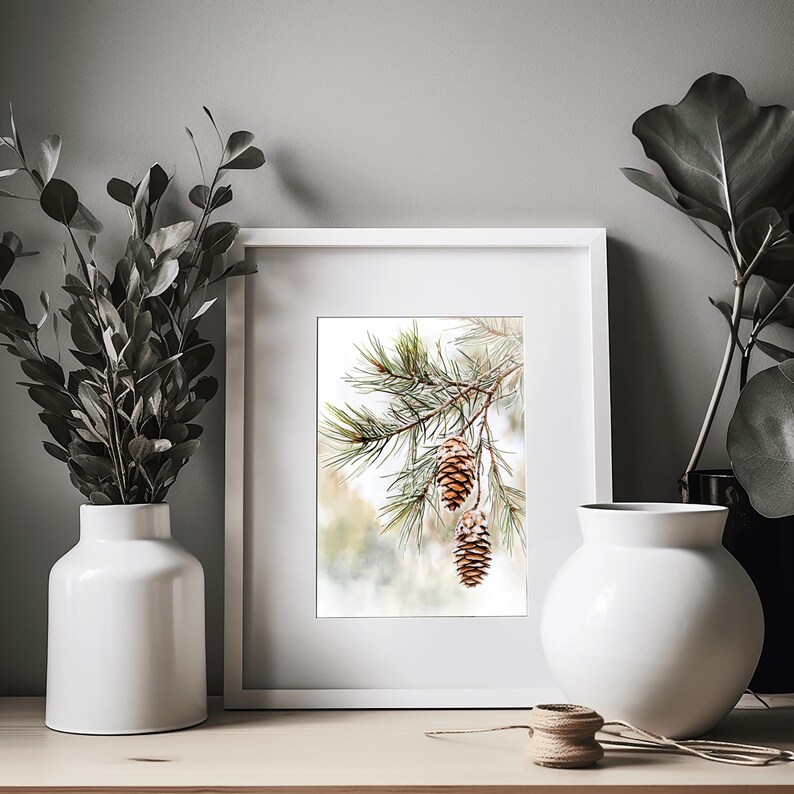May include: A framed print of a watercolor painting featuring a close-up of a pine branch with three pine cones. The background is a soft, blurred watercolor wash of green and brown.