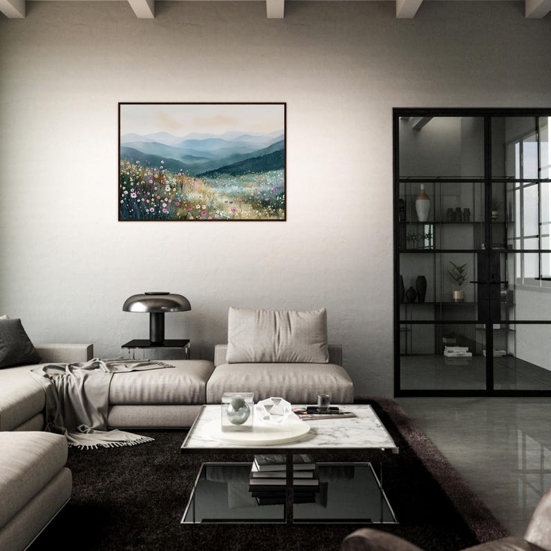 May include: A rectangular landscape painting in a thin black frame hangs on a wall. The artwork depicts a field of colorful wildflowers in the foreground with rolling blue and green mountain ranges fading into a soft, hazy sky in the distance.