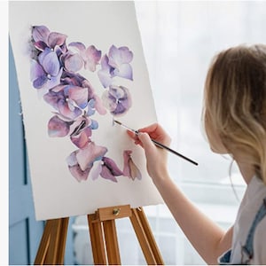 May include: A woman paints a watercolor floral design on a canvas on an easel. The flowers are purple, pink, and white.