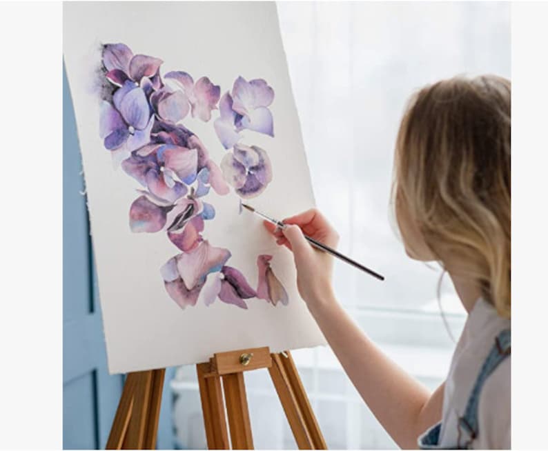 May include: A woman paints a watercolor floral design on a canvas on an easel. The flowers are purple, pink, and white.