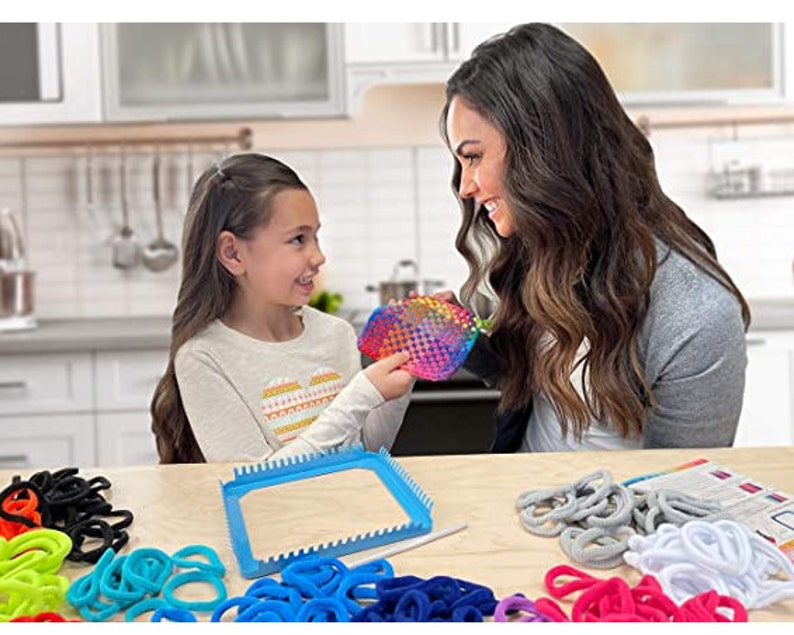 Puede incluir: Una ni&ntilde;a y una mujer est&aacute;n sentadas en una mesa haciendo pulseras de loom arco&iacute;ris. Est&aacute;n usando un telar y gomas el&aacute;sticas de colores. El telar es azul y tiene forma cuadrada. Hay muchas gomas el&aacute;sticas de colores en la mesa.