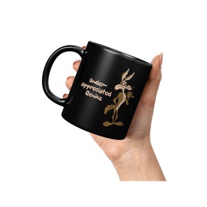 A black coffee mug with a cartoon Wile E Coyote in brown and tan colors. The coyote is standing with its right paw raised. The text on the mug reads 'Under-appreciated Genius'.