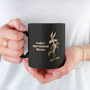 A black coffee mug with a cartoon Wile E Coyote in brown and tan colors. The coyote is standing with its right paw raised. The text on the mug reads 'Under-appreciated Genius'.