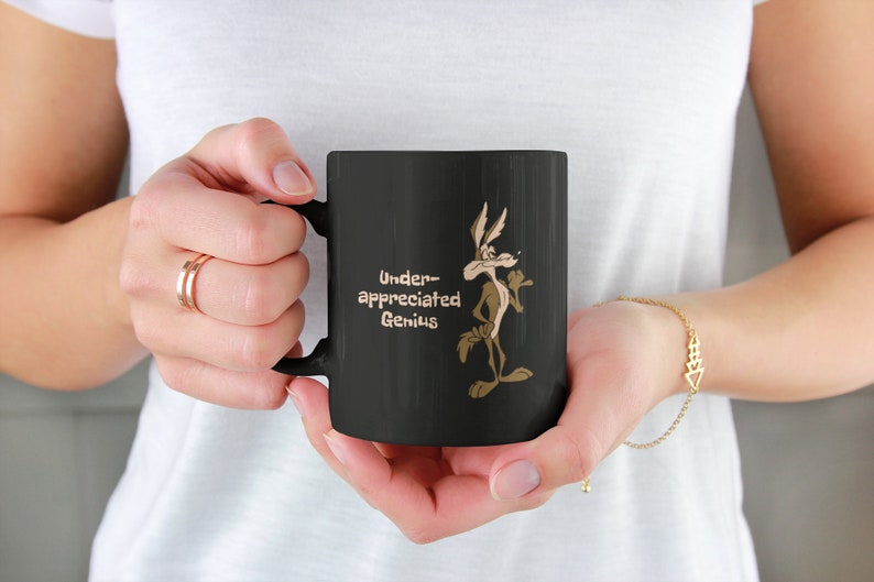 A black coffee mug with a cartoon Wile E Coyote in brown and tan colors. The coyote is standing with its right paw raised. The text on the mug reads 'Under-appreciated Genius'.