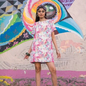 Może przedstawiać: Kobieta w różowo-białej sukience z tie-dye, krótkimi rękawami i czarną opaską na głowie. Stoi przed kolorowym muralem z dużym wzorem oka. Ma na sobie czarne sznurowane buty.