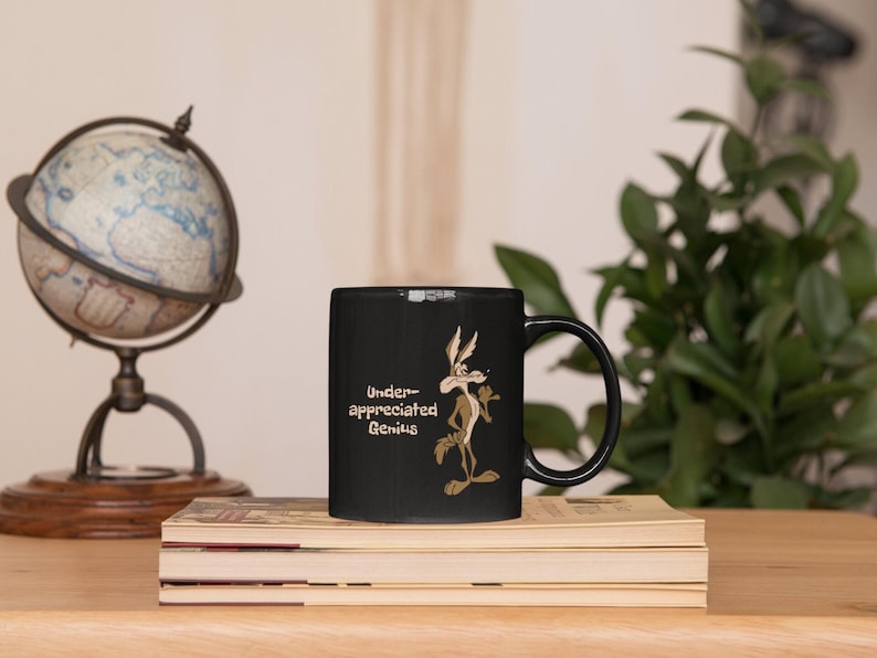 A black coffee mug with a cartoon Wile E Coyote in brown and tan colors. The coyote is standing with its right paw raised. The text on the mug reads 'Under-appreciated Genius'.