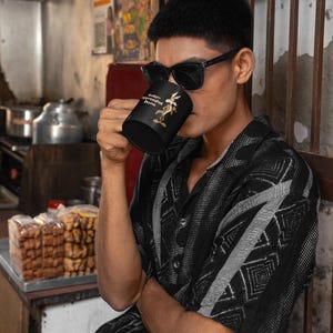 A black coffee mug with a cartoon Wile E Coyote in brown and tan colors. The coyote is standing with its right paw raised. The text on the mug reads 'Under-appreciated Genius'.