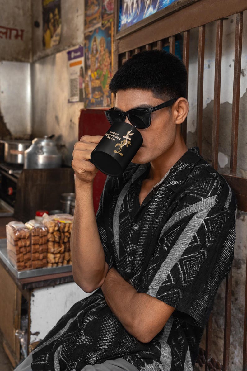 A black coffee mug with a cartoon Wile E Coyote in brown and tan colors. The coyote is standing with its right paw raised. The text on the mug reads 'Under-appreciated Genius'.