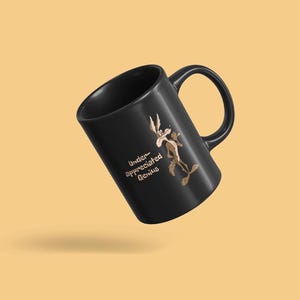 A black coffee mug with a cartoon Wile E Coyote in brown and tan colors. The coyote is standing with its right paw raised. The text on the mug reads 'Under-appreciated Genius'.