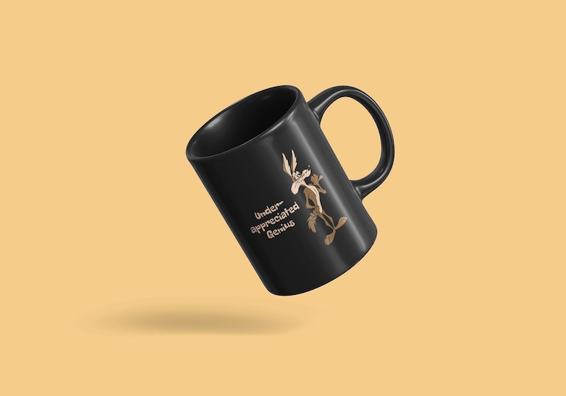 A black coffee mug with a cartoon Wile E Coyote in brown and tan colors. The coyote is standing with its right paw raised. The text on the mug reads 'Under-appreciated Genius'.
