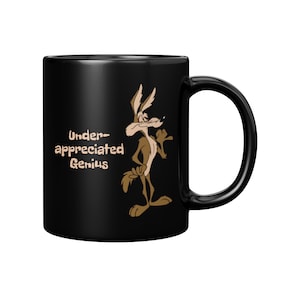 A black coffee mug with a cartoon Wile E Coyote in brown and tan colors. The coyote is standing with its right paw raised. The text on the mug reads 'Under-appreciated Genius'.
