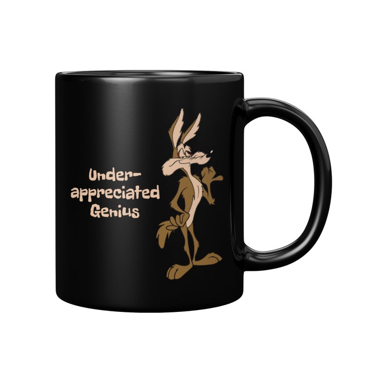 A black coffee mug with a cartoon Wile E Coyote in brown and tan colors. The coyote is standing with its right paw raised. The text on the mug reads 'Under-appreciated Genius'.