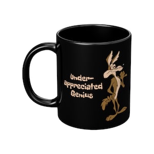 A black coffee mug with a cartoon Wile E Coyote in brown and tan colors. The coyote is standing with its right paw raised. The text on the mug reads 'Under-appreciated Genius'.