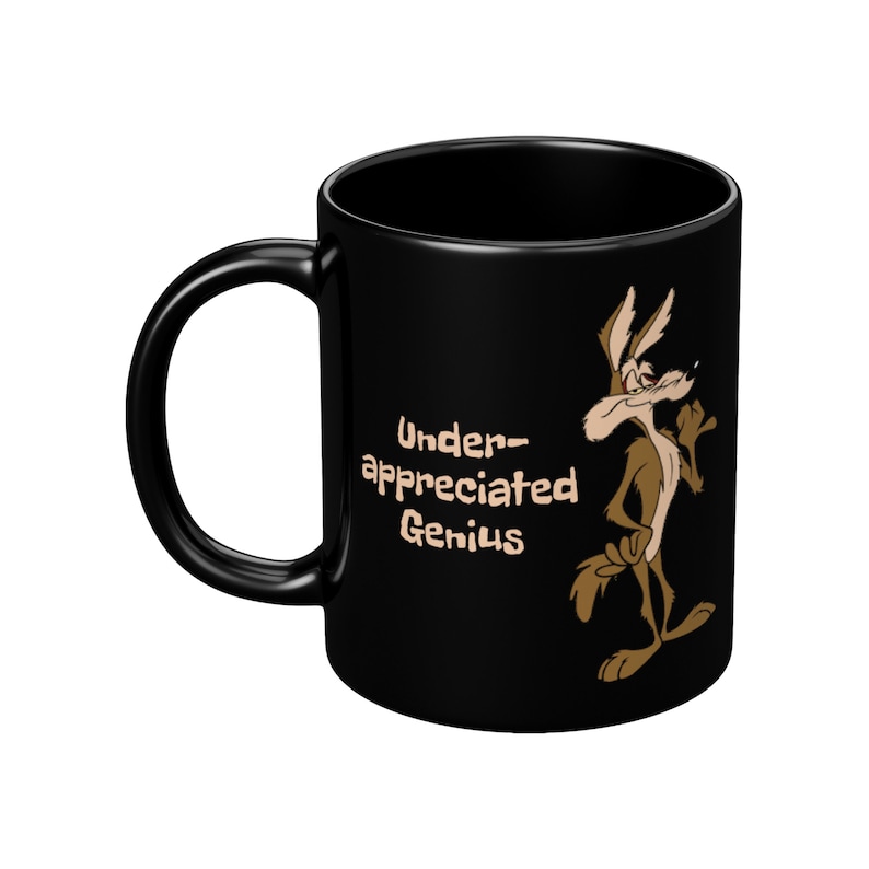 A black coffee mug with a cartoon Wile E Coyote in brown and tan colors. The coyote is standing with its right paw raised. The text on the mug reads 'Under-appreciated Genius'.