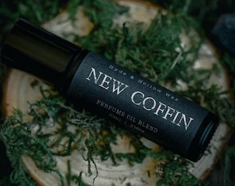 May include: A black roller bottle with a label that reads "Hyde & Hollow Wax New Coffin Perfume Oil Blend 10ml | .33oz". The bottle is sitting on a wooden disc surrounded by green moss.