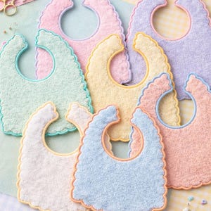 Op de afbeelding: Een verzameling pastelkleurige baby slabbetjes met geschulpte randen en contrasterende afwerking. De slabbetjes zijn in verschillende kleuren, waaronder roze, geel, blauw, groen en lavendel. Naaibenodigdheden, zoals garen en scharen, zijn zichtbaar.