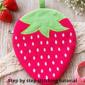May include: A pink strawberry-shaped coin pouch with a green stem and white seed details. The pouch has a zipper closure and a small green loop. Text on the image reads "Strawberry Zipper Purse Embroidery Design In The Hoop Coin Pouch" and "Step by step stitching tutorial included."