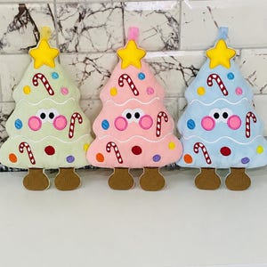 Pode incluir: Três enfeites de árvore de Natal de pelúcia nas cores verde-claro, rosa-claro e azul-claro. Cada árvore possui uma estrela amarela no topo, dois olhos brancos com pupilas pretas, bochechas rosas e pés marrons, decorados com bengalas doces.
