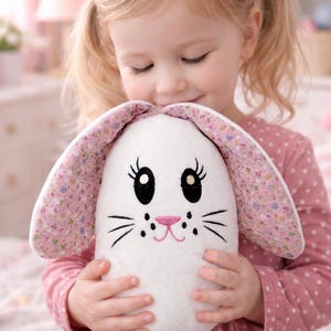 Peut inclure: Une peluche blanche en forme de lapin avec des oreilles à motifs floraux roses et un nez rose. Le lapin a des yeux, des moustaches et une bouche souriante brodés en noir. Le jouet est tenu par un enfant portant une robe rose à pois.