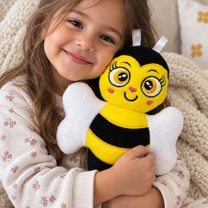 Könnte beinhalten: Ein Plüsch-Bienen-Spielzeug mit einem gelb-schwarz gestreiften Körper, großen gestickten Augen mit langen Wimpern, rosa Wangen, einer kleinen herzförmigen Nase und weißen Flügeln. Zwei kleine weiße Stoffantennen sind oben am schwarzen Kopf befestigt.