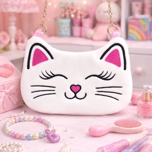 May include: White cat-shaped handbag with a gold chain strap. The bag features black whiskers, closed eyes, and pink accents on the ears and heart-shaped nose. The bag is on a pink surface with other accessories.