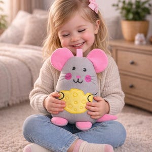 Puede incluir: Un peluche gris con forma de ratón con orejas, pies y cola rosas, y un detalle de queso amarillo. El juguete lo sostiene un niño pequeño que lleva vaqueros azules y un jersey color crema. El niño está sentado en una alfombra clara.