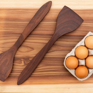 May include: Two wooden spatulas, one with a pointed end and the other with a rounded end, are laid out on a wooden chopping board. A carton of six brown eggs sits on the board.